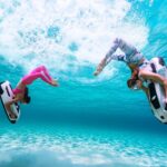 Two people riding jet skis underwater with waves above.