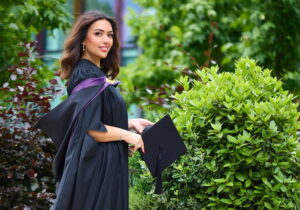 A smiling graduate in a black gown holding a cap outdoors.