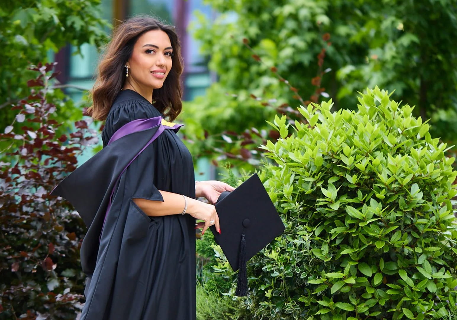 A smiling graduate in a black gown holding a cap outdoors.