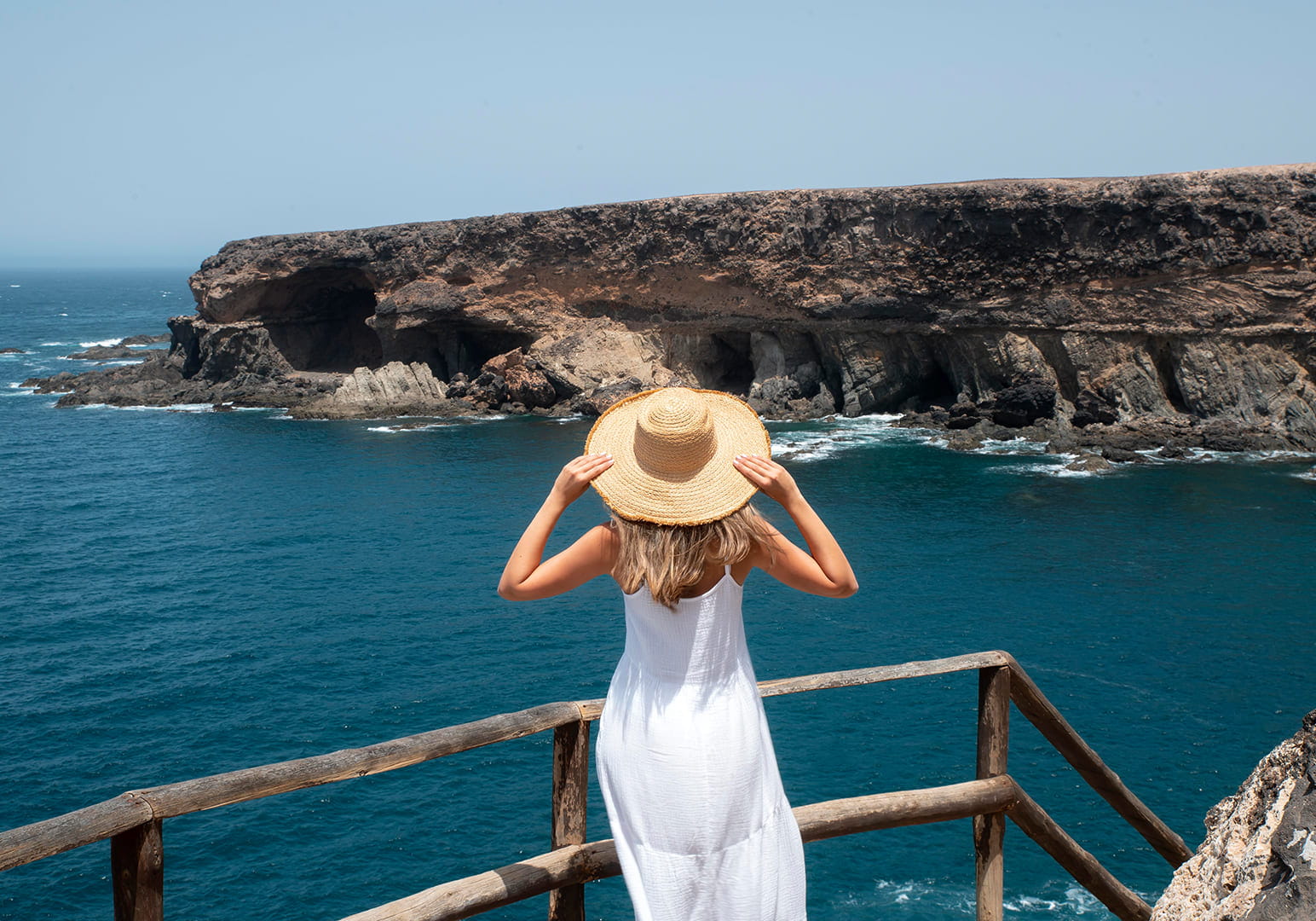 Woman in white dress and sunhat overlooking rocky coastline and ocean.